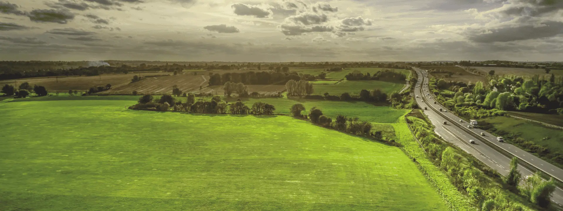 Aerial view of a green landscape featuring a highway on the right. Sunlight breaks through the clouds, illuminating fields and trees in the background.
