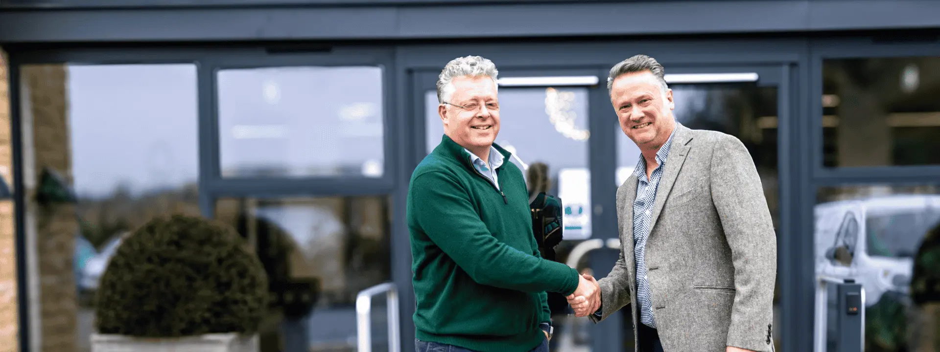 Two men shaking hands outside a modern building entrance, one wearing a green sweater and the other in a gray blazer, both smiling.