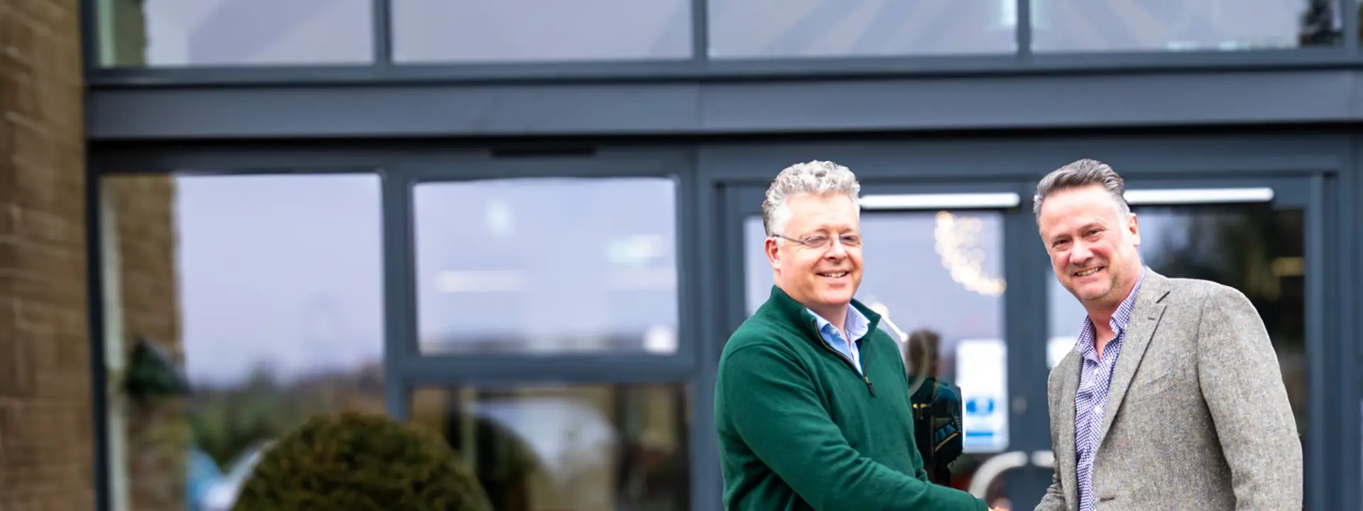 Two men shaking hands in front of a modern building entrance, with glass doors and a decorative archway. One man wears a green sweater and the other a gray blazer, both are smiling.