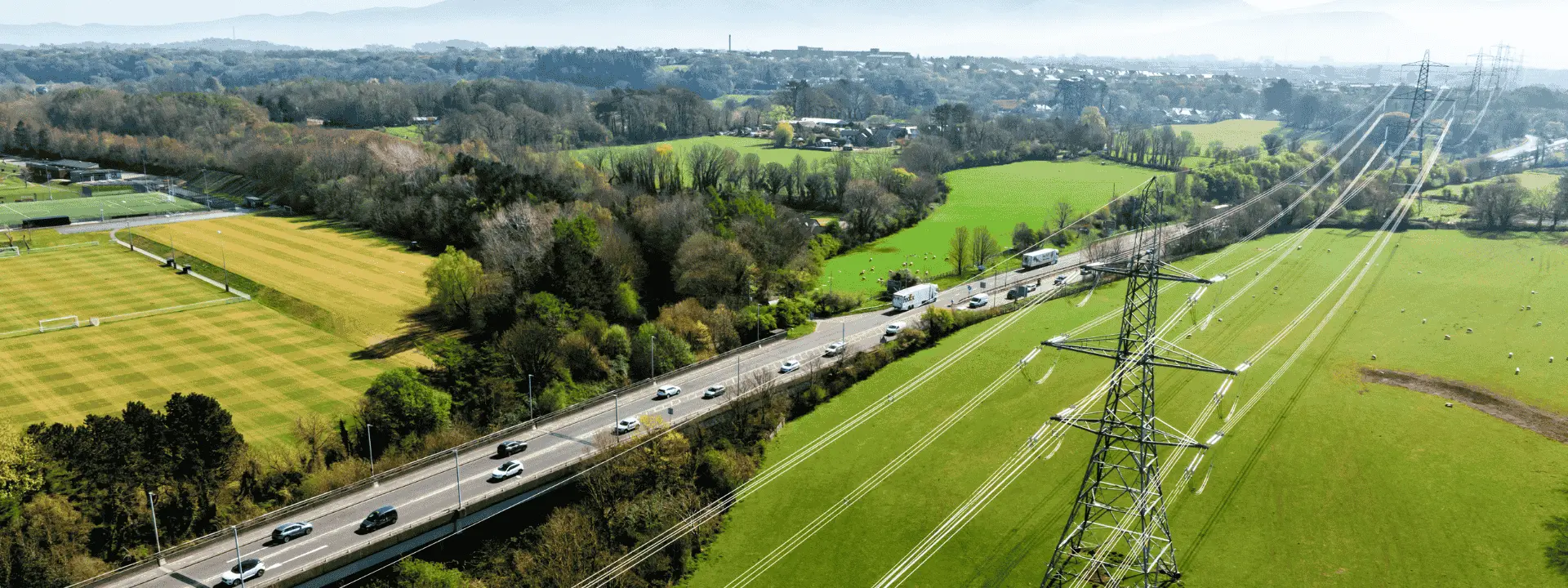 An aerial view of a rural landscape featuring a highway lined with vehicles, green fields, and utility poles. In the background, there are mountains and a clear blue sky.