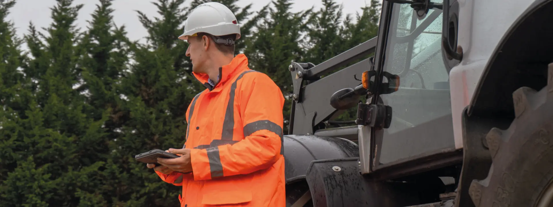 A construction worker in an orange safety jacket and white hard hat stands beside a large machinery, holding a tablet while looking at it. Green trees are in the background.