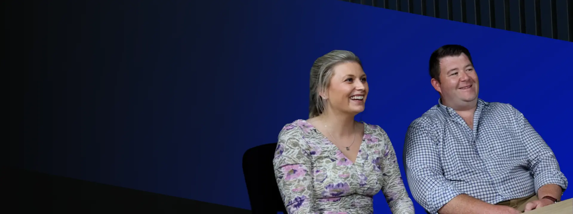 Two individuals sitting at a wooden table in a modern office setting, with a blue and dark background. The woman wears a floral dress and smiles, while the man, in a checked shirt, is seated beside her.