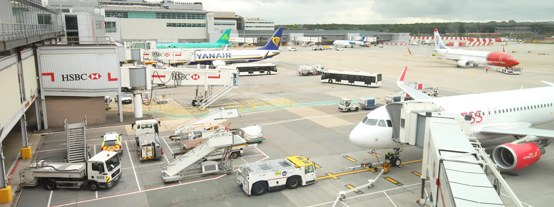 An airport scene showing multiple airplanes parked at gates, including a Yanair flight, and ground service vehicles assisting with luggage at a busy terminal.