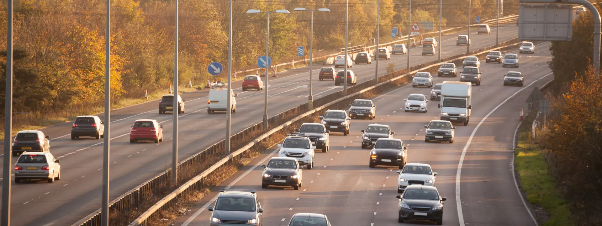 A view of a multi-lane highway with several vehicles, including cars and a truck, traveling in both directions. The scene is set during sunset, with trees and fields visible in the background.