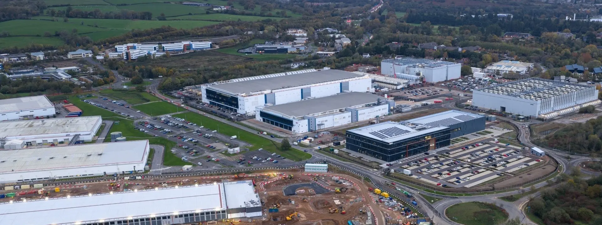 An aerial view of a commercial area featuring several large industrial buildings, parking lots, and areas under construction. The landscape includes green fields and scattered trees, with hills in the background.