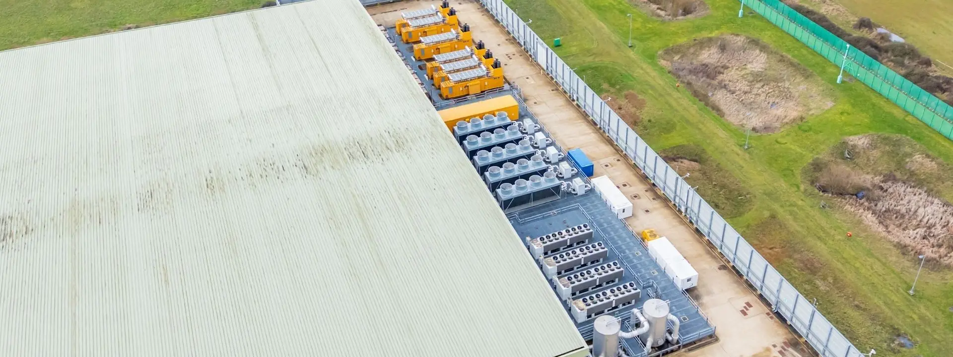 An aerial view of a large industrial building with a green roof. The site features several large machinery units and containers arranged along one side, surrounded by a fenced area with grassy fields and land in the background.