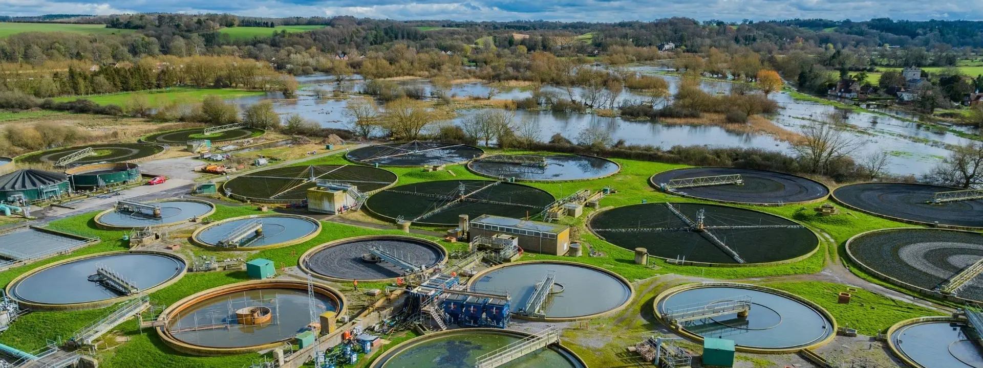 An aerial view of a water treatment facility featuring numerous large, circular tanks arranged across a green landscape. Surrounding the facility, there are areas of grass and trees, with a river visible in the background.