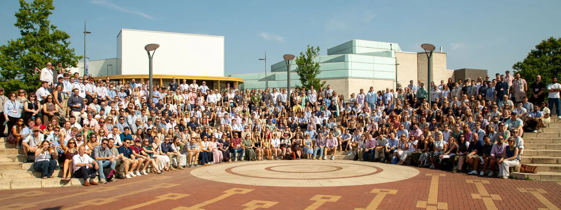 A large group of people gathered outdoors on a paved area featuring a circular design. They are arranged in rows on steps, with buildings and trees in the background under a clear sky.
