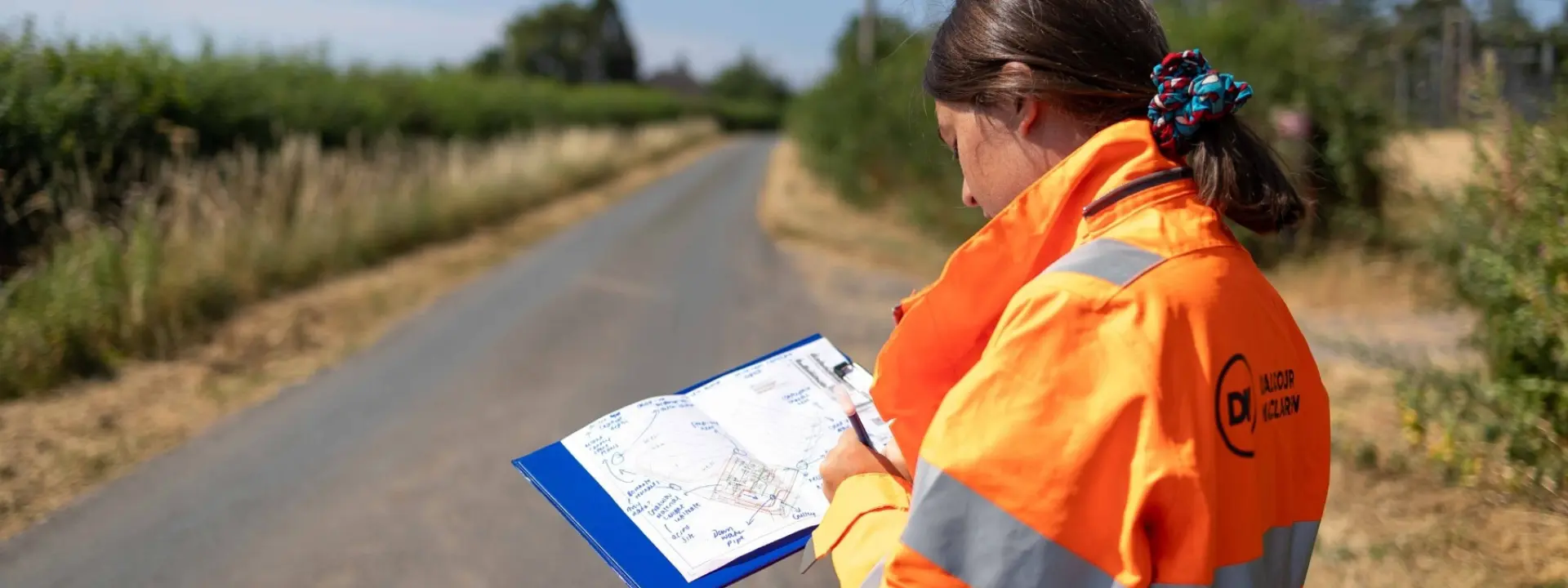 A worker in a bright orange safety jacket studies a clipboard with maps while standing on a rural road.