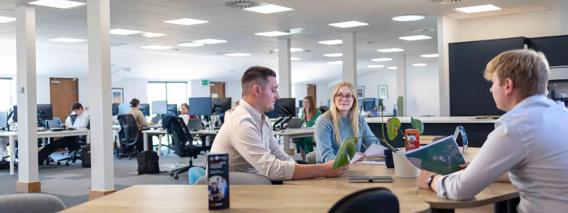 Three office workers sit around a wooden table in an open-plan office, with desks, computers, and large wall monitors in the background.
