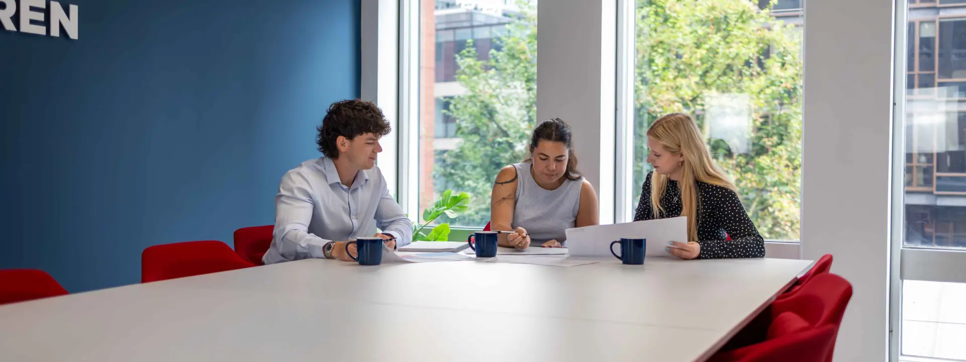 Three coworkers sit at a large conference table reviewing papers in a bright meeting room with red chairs and large windows.