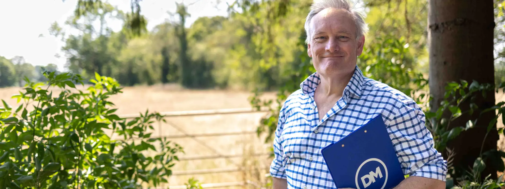 A man in a blue checkered shirt holds a blue DM folder while standing outdoors beside a tree and green shrubs, with a sunlit field behind him.