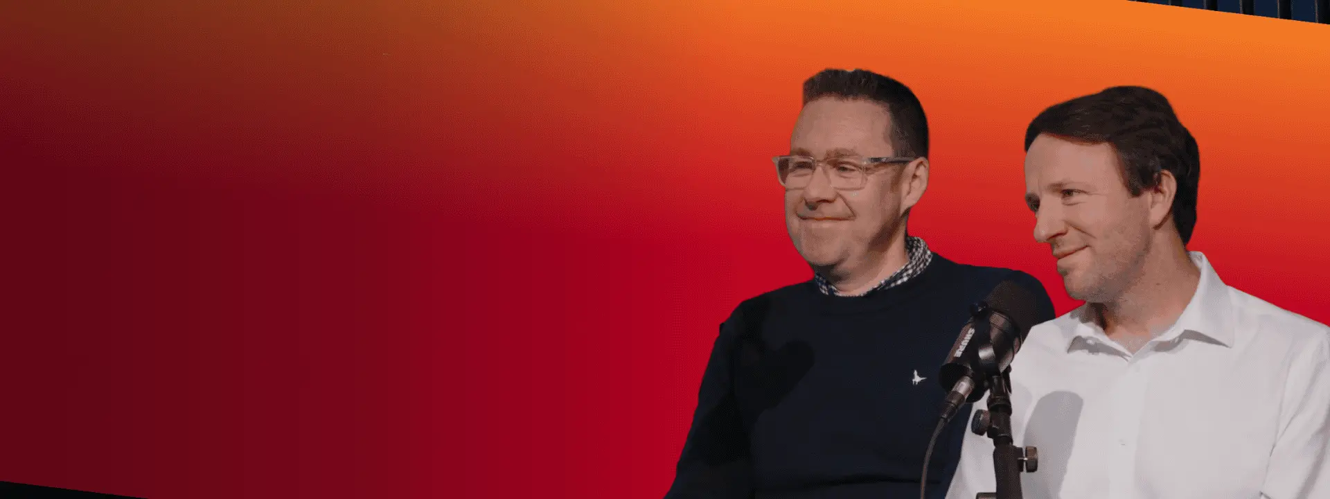 Two men stand beside a microphone in front of a large red-to-orange gradient screen, with dark vertical slats visible above.