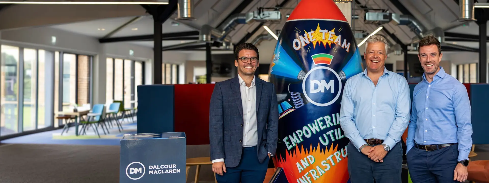 Three men stand beside a large rocket-shaped display reading “ONE TEAM,” “DM,” and “Empowering Utility and Infrastructure” in a modern office space.