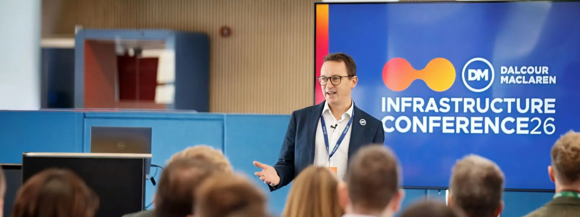 A man in a suit speaks at an infrastructure conference, standing beside a blue presentation screen while an audience sits in the foreground.