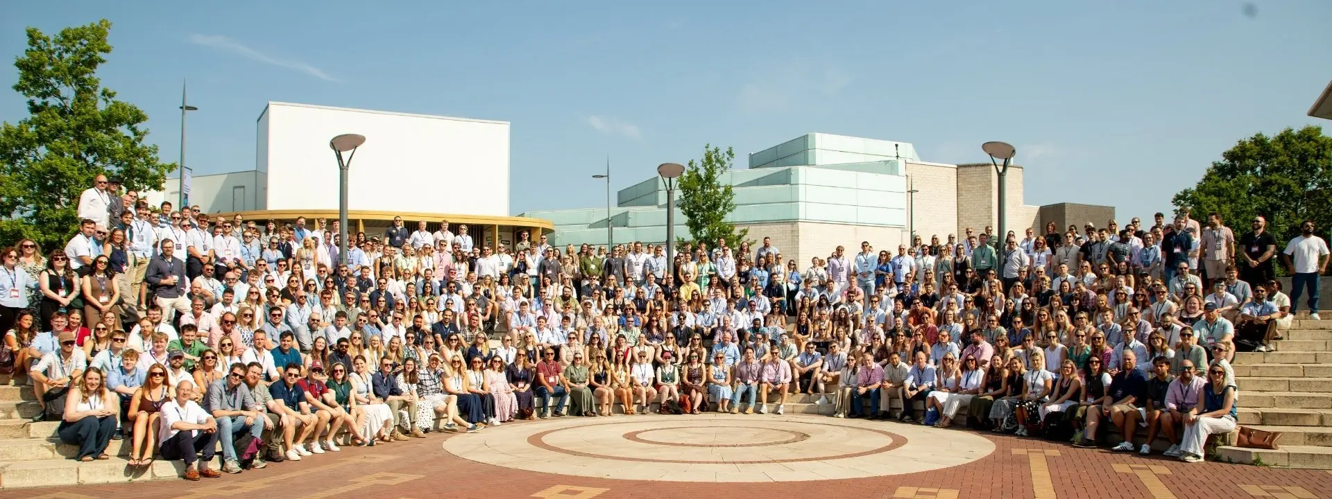 A large group of people poses on a tiled plaza in front of modern buildings under a clear blue sky.