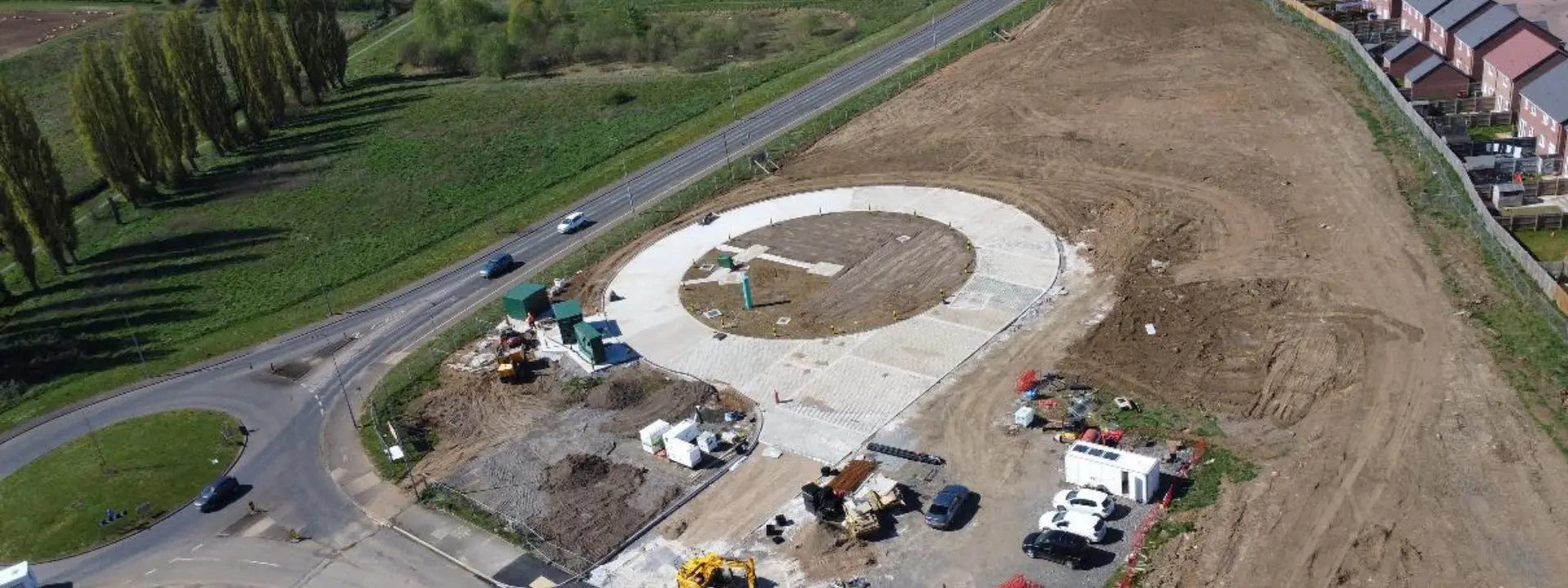 An aerial view of a construction site featuring a circular concrete foundation for a roundabout, surrounded by cleared land and vehicles. In the background, residential houses are visible alongside green fields.