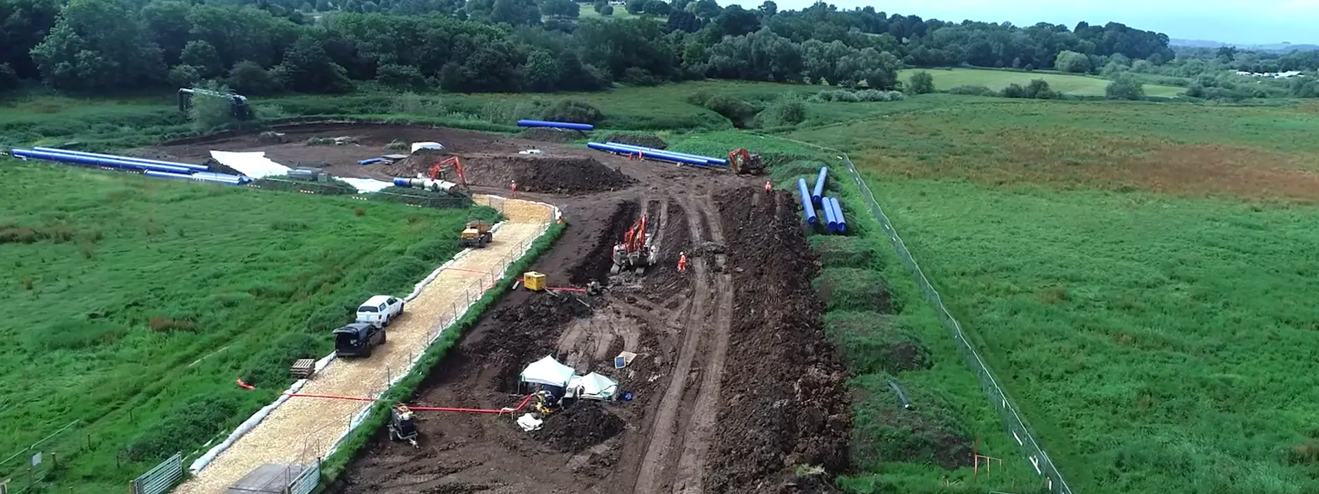 An aerial view of a construction site with heavy machinery and vehicles working on a dirt road. The area is surrounded by green fields and trees in the background under a cloudy sky.