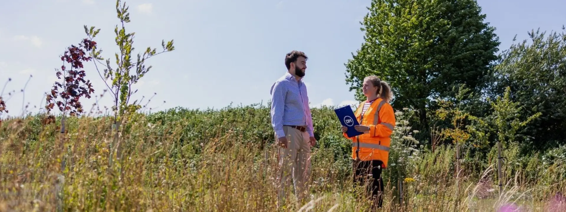 A man and a woman in an orange safety jacket stand in a grassy field, with the woman holding a blue clipboard and trees in the background.