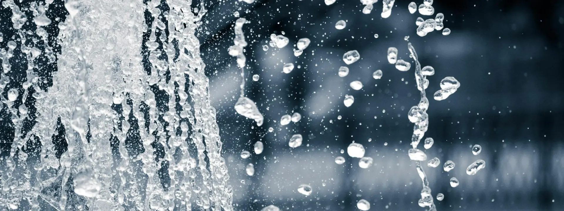Close-up of water droplets splashing from a fountain against a blurred background. The image captures the motion and texture of the water as it cascades.