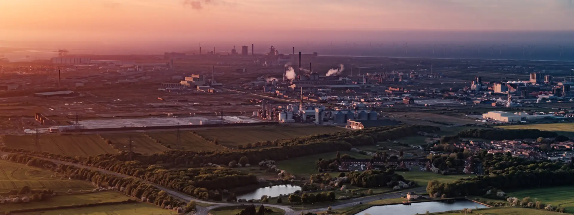A panoramic view of an industrial landscape during sunset, featuring factories and smokestacks in the background, surrounded by green fields and bodies of water.