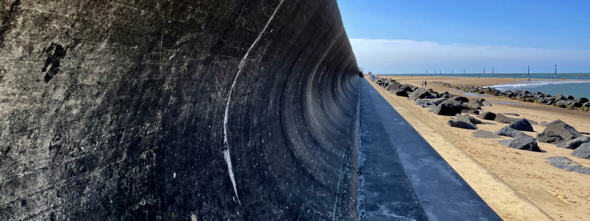A curved concrete wall along a beach, with a coastline visible in the background. The sky is clear and blue, and a line of rocks runs along the sand beside the water.