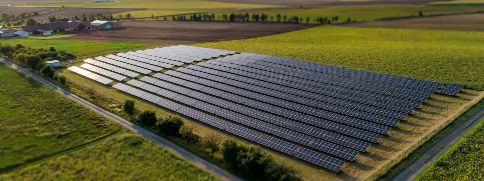 An aerial view of a solar farm with rows of solar panels arranged in a grid pattern, surrounded by green fields and farmland. A dirt road leads around the solar installation.