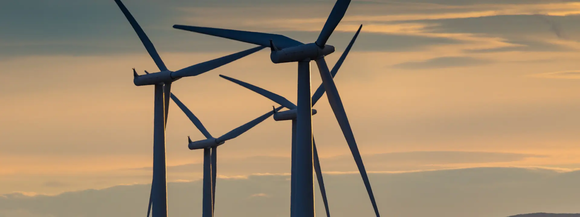 Three wind turbines silhouetted against a colorful sunset, with a vast open landscape in the background.