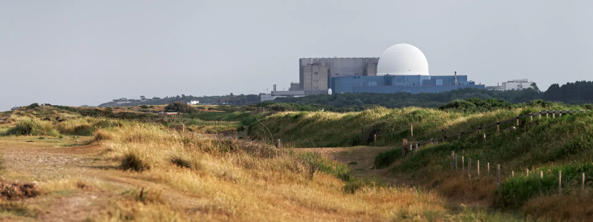A nuclear power plant with a large white dome in the background, surrounded by grassy terrain and a sandy path leading through the landscape.