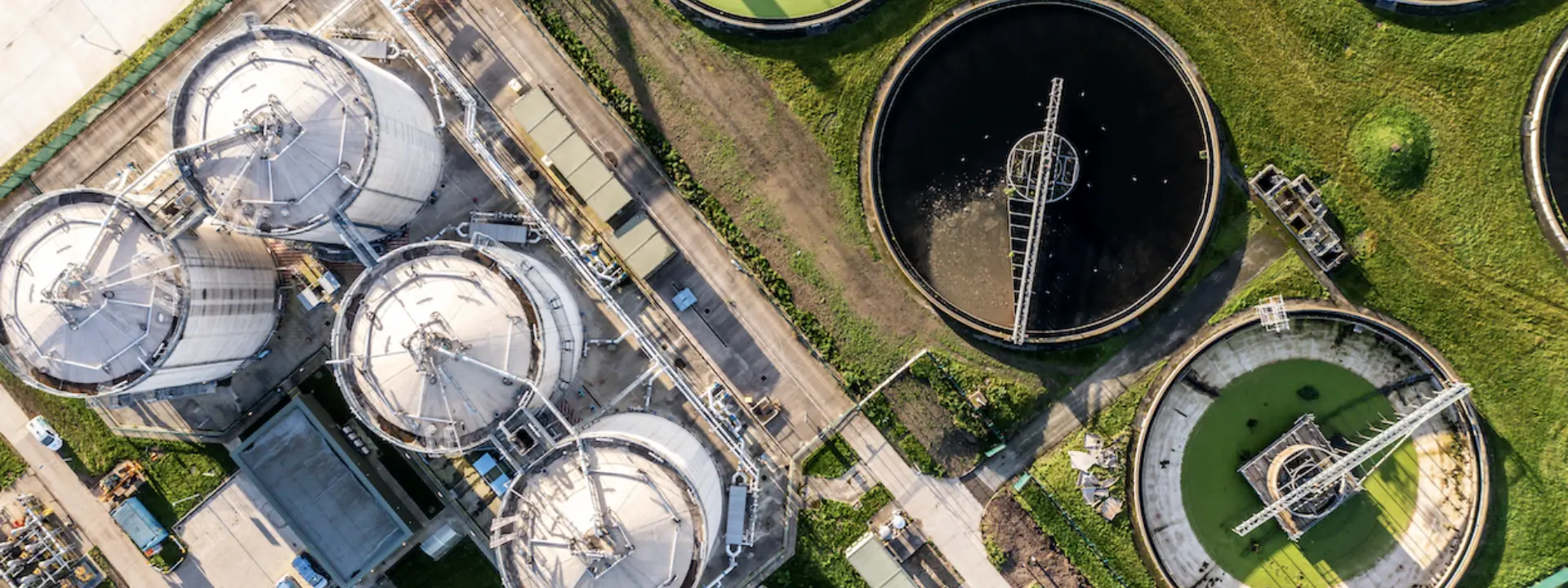 An aerial view of a wastewater treatment facility featuring several large, circular tanks filled with green and dark water. Nearby structures and pipes are also visible on the grass-covered ground.
