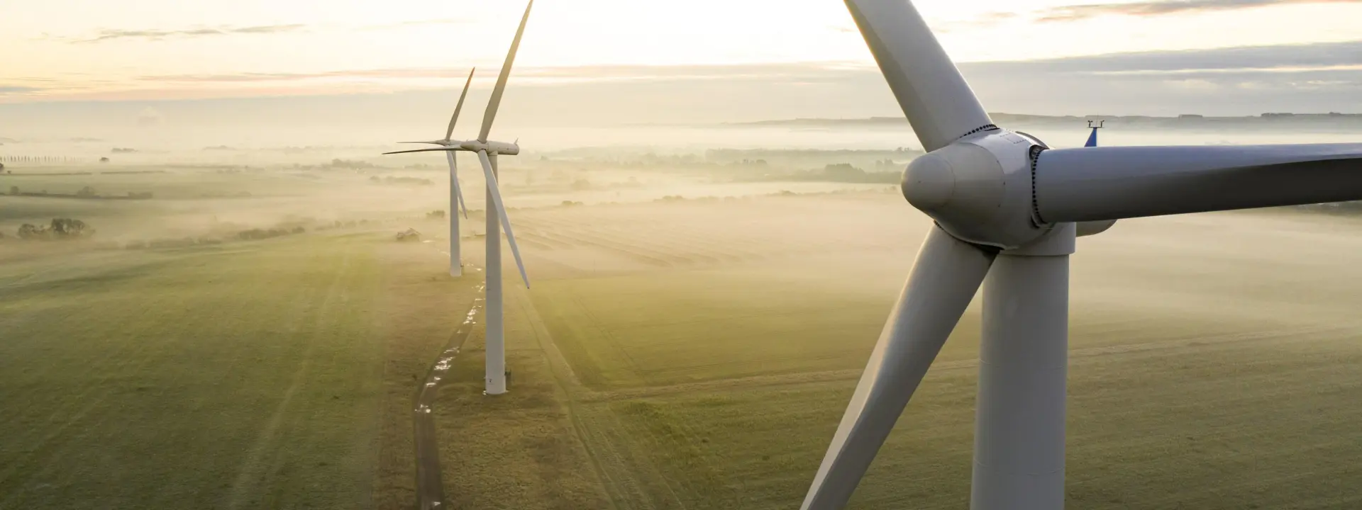 Aerial view of several wind turbines in a green landscape during early morning light, with mist covering the ground and a winding path visible in the foreground.