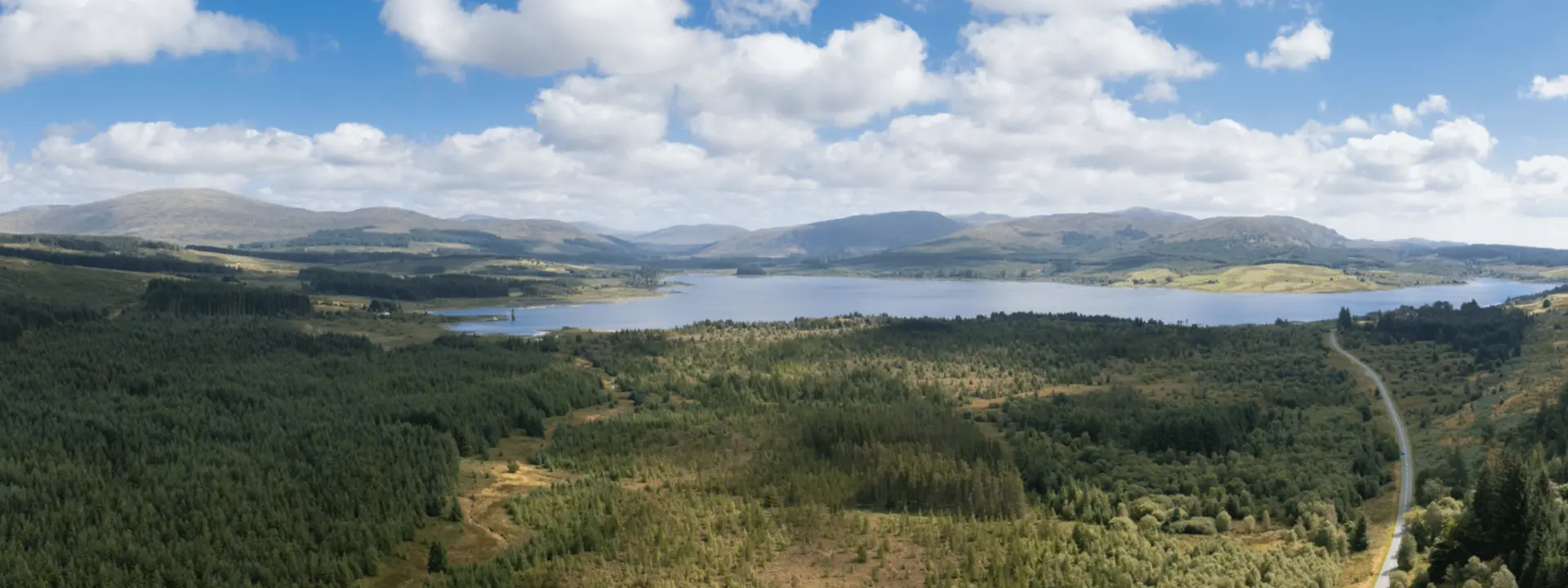 A panoramic view of a lake surrounded by green forests and rolling hills under a partly cloudy sky. A winding road is visible on the right side of the image.