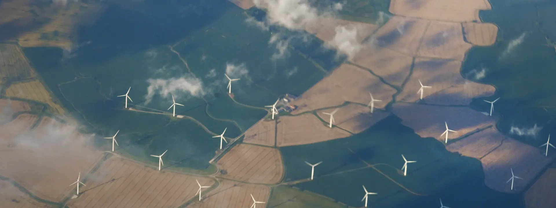 Aerial view of farmland with wind turbines scattered across the fields, under a partly cloudy sky. The landscape features patches of different colors indicating various crops.
