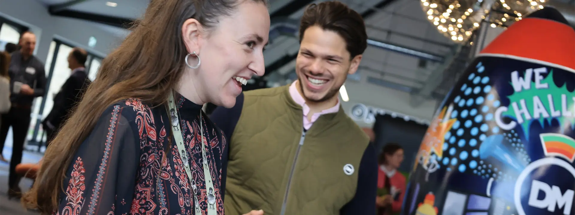 Two smiling individuals engaged in conversation at a lively event. The woman, wearing a patterned blouse, holds a small object while the man, dressed in a green vest, appears amused. A vibrant display with the text 'WE CHALLENGE' is visible in the background.