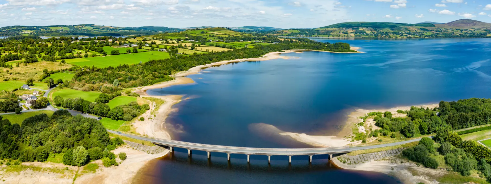Aerial view of a river with a bridge spanning across it, surrounded by lush green fields and rolling hills under a bright blue sky with fluffy clouds.