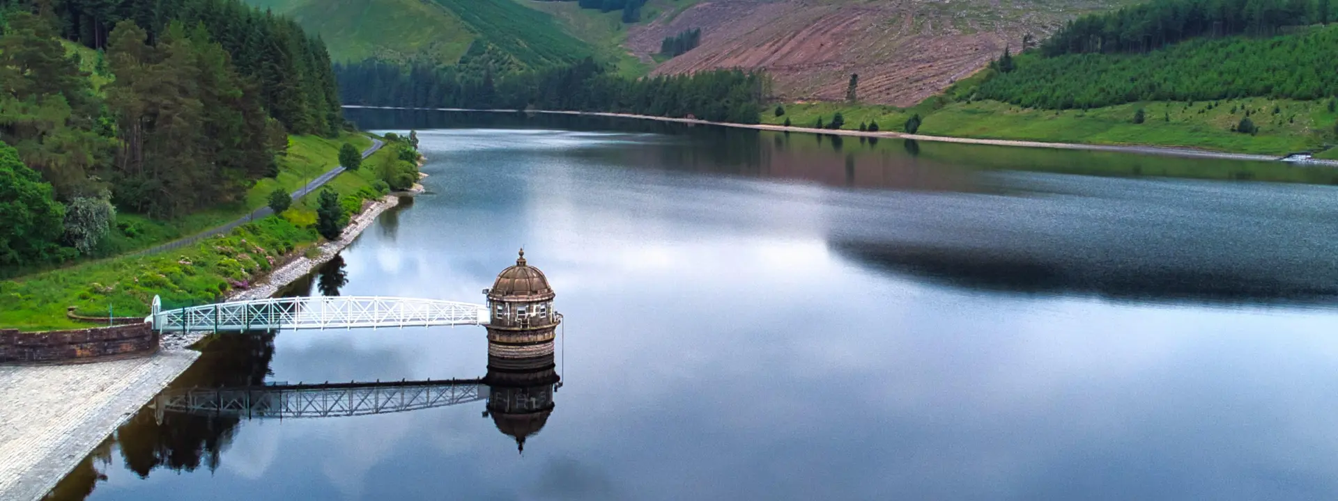 A scenic view of a calm lake surrounded by green hills, with a small decorative structure at the water's edge and a walking path along the shore. The sky is partly cloudy, reflecting on the water's surface.