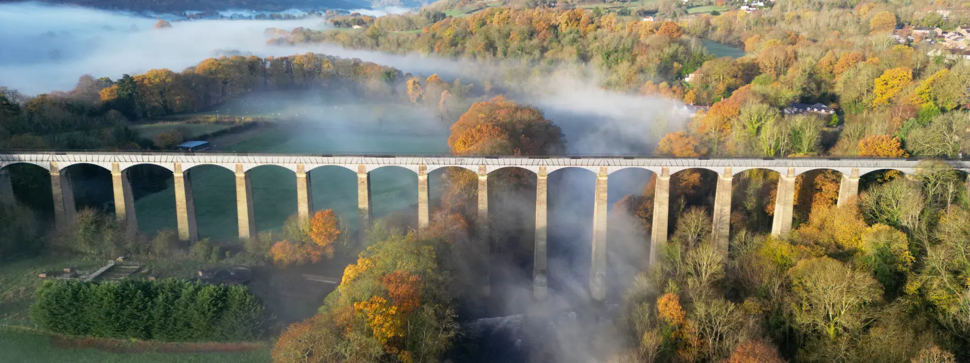 Aerial view of a stone viaduct with multiple arches spanning over a misty landscape filled with autumn-colored trees and low-lying fog.
