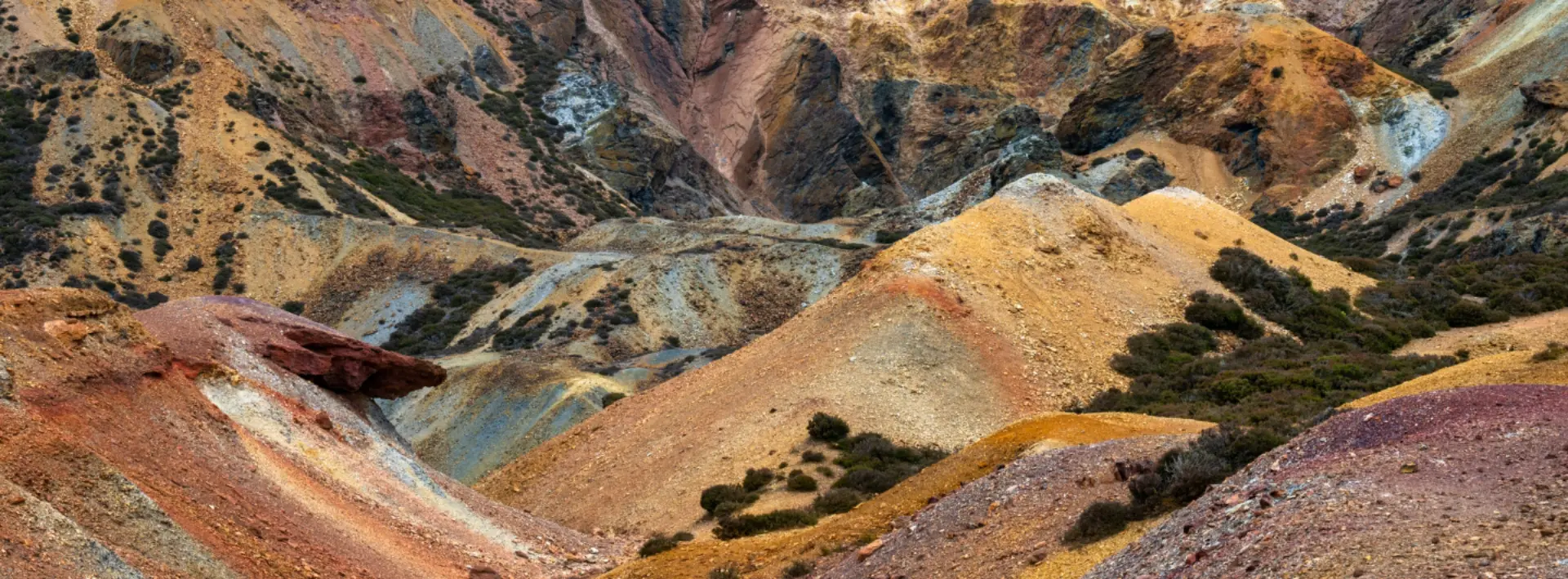 Colorful mountain range with varying shades of orange, yellow, and brown. The landscape features smooth hills and rocky slopes, with patches of green vegetation scattered throughout.