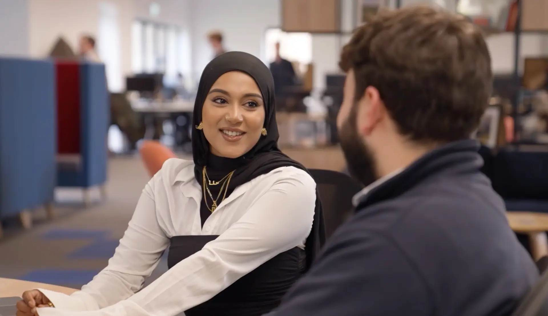 A woman in a white blouse and black headscarf sits at a desk, smiling while engaging in conversation with a man in a dark sweater, both in a modern office environment.
