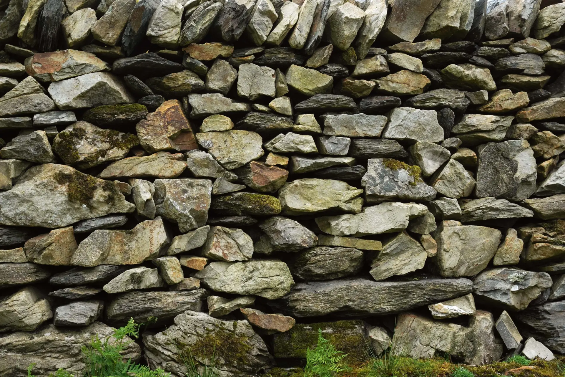 A close-up view of a dry-stone wall made up of various sizes and shapes of gray and tan stones, with patches of moss and grass visible at the base.