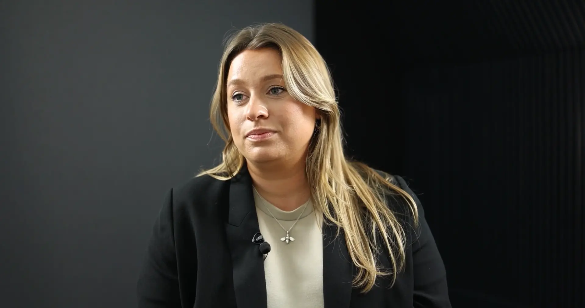 A woman with long blonde hair, wearing a black blazer over a light top, sitting in front of a dark background, looking off to the side.