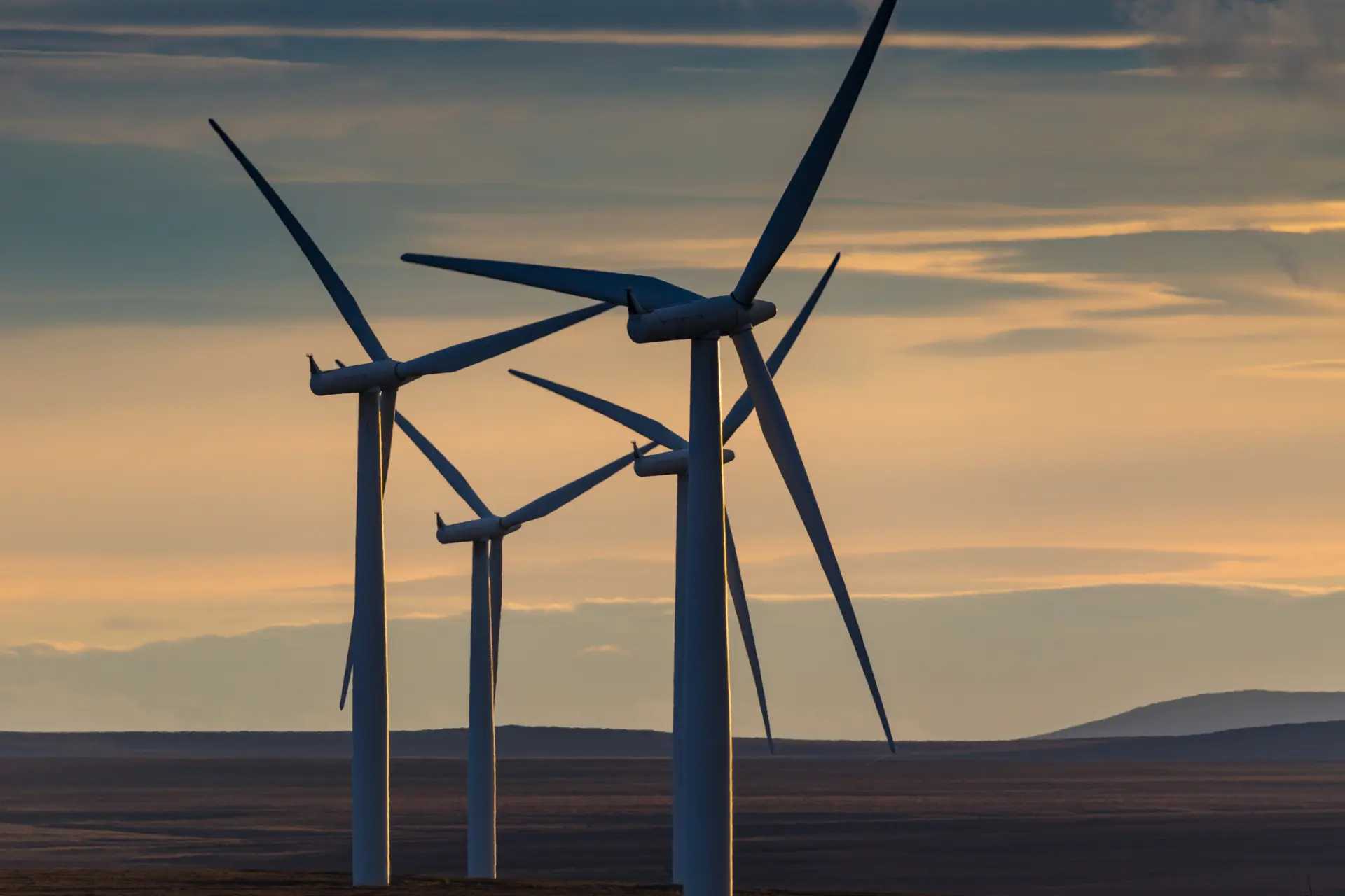 Three wind turbines silhouetted against a colorful sunset, with a vast open landscape in the background.
