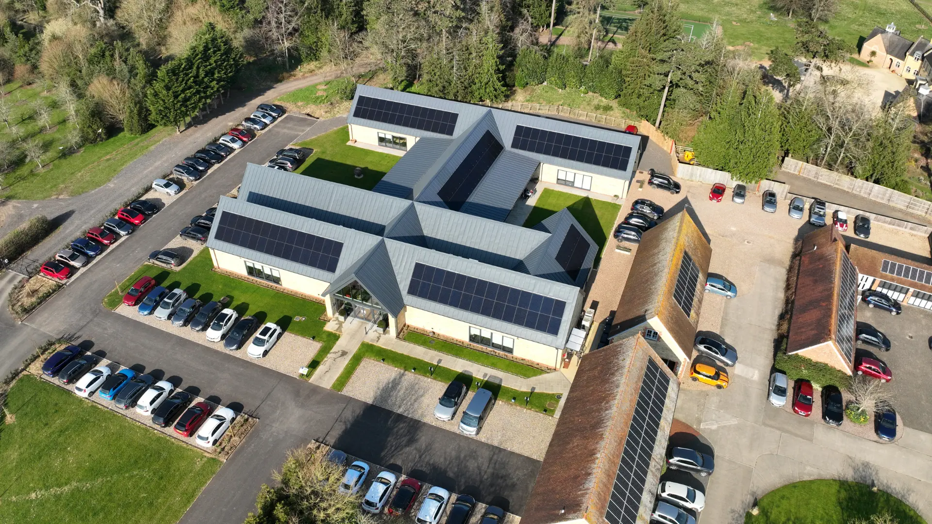 Aerial view of a large building with solar panels on the roof, surrounded by green grass. Multiple parked cars are visible in the parking lot, along with neighboring buildings with solar installations, set in a natural landscape.