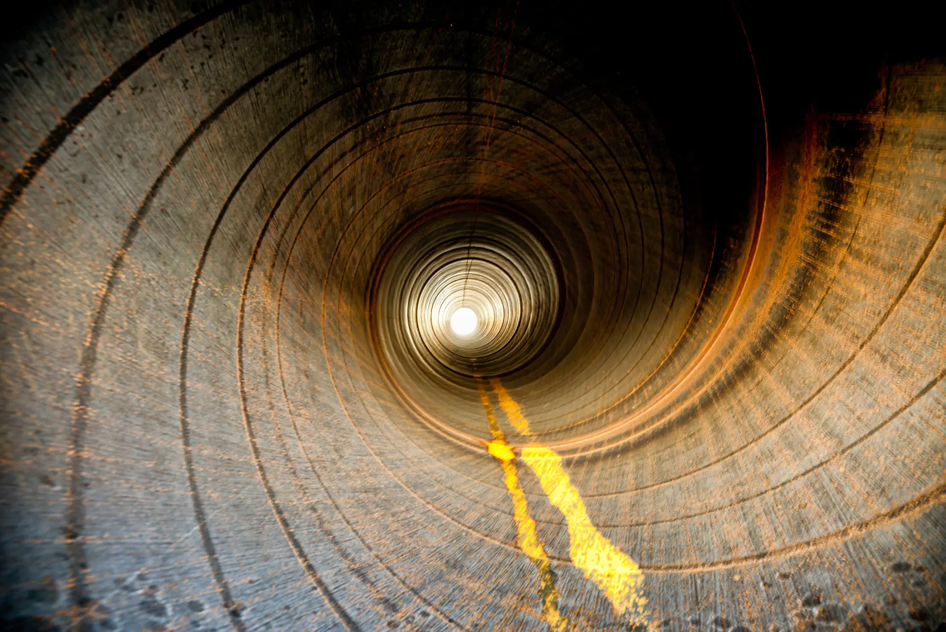 View inside a large, cylindrical pipe with a bright light at the far end, showing a textured surface and a faint yellow line running along the center.