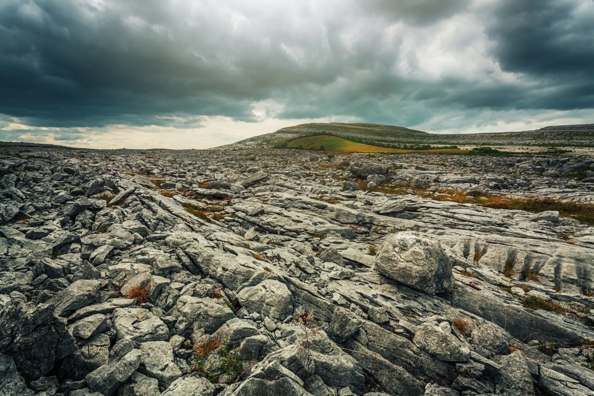 A rocky landscape with large stone slabs and patches of grass under a cloudy sky. Rolling hills are visible in the background, and the terrain appears rugged and uneven.