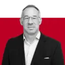 A man wearing glasses and a black blazer over a striped shirt, posing against a red background.