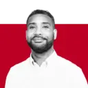 A smiling man with facial hair wearing a white collared shirt, against a background featuring black and red colors.