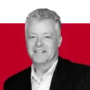A smiling man with gray hair wearing a white shirt and black blazer, set against a background featuring solid green and red sections.