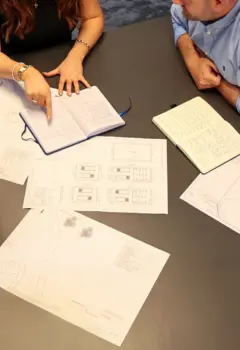 A close-up view of a meeting table with architectural plans and sketches spread out. Two individuals are engaged in discussion, one pointing at an open notebook while the other holds a closed notebook. The setting appears to be a collaborative workspace.