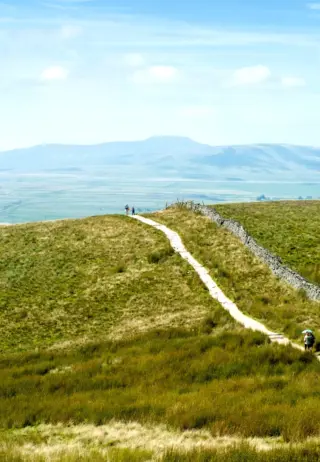 A scenic view of rolling hills under a blue sky, with a winding dirt path leading through green grass. A few people are walking along the path, enjoying the expansive landscape in the background.